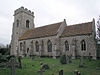 Eine steinerne Kirche mit einem massiven Kirchturm auf der linken Seite; das Kirchenschiff hat drei Fenster und der niedrigere Altarraum hat zwei Fenster.