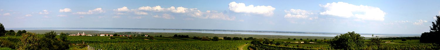 Panoramabild des Neusiedler Sees von Westen in den Weinbergen über Rust aufgenommen. Der Standort gewährt einen Blick bis weit hinein in die Pannonische Tiefebene. Links im Vordergrund die Stadt Rust, rechts im Vordergrund die Seebühne von Mörbisch