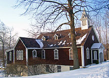 Ein mit dunkelbraunem Holz verkleidetes Gebäude mit spitzem Dach und einem kleinen Türmchen am rechten Ende. Schnee liegt auf dem Boden in der Umgebung, Reste auf dem Dach. Ein großer blattfreier Baum steht vor dem Vauwerk