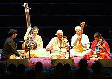 Eine Gruppe von fünf Männern und Frauen sitzt auf einer Plattform, zwei spielen langhalsige Lauten, zwei andere spielen Sreichinstrumente und einer stützt seine Hände neben Trommeln auf.