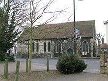 Photographie einer Kirche, ein Busch im Bildvordergrund. Eine Straße verläuft vor dem Kirchengebäude.