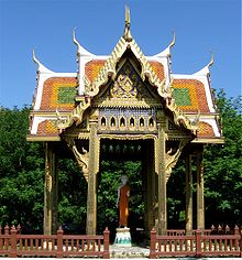 ein hoch aufgeständertes Holzdach mit geschnitzten Giebeln und bunten Dachziegeln über einer Buddha-Statue