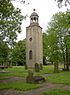 Ein schlanker steinerner Kirchturm in einem Kirchhof mit zwei Fenstern und einer Uhr, darüber befindet sich eine Kuppel.